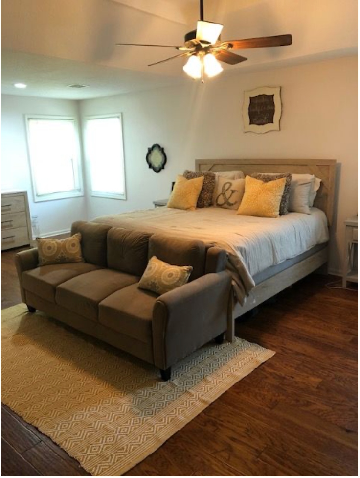 Staged master bedroom featuring cozy linen bedding, yellow textured accent pillows, and an inviting sofa at the foot of the bed